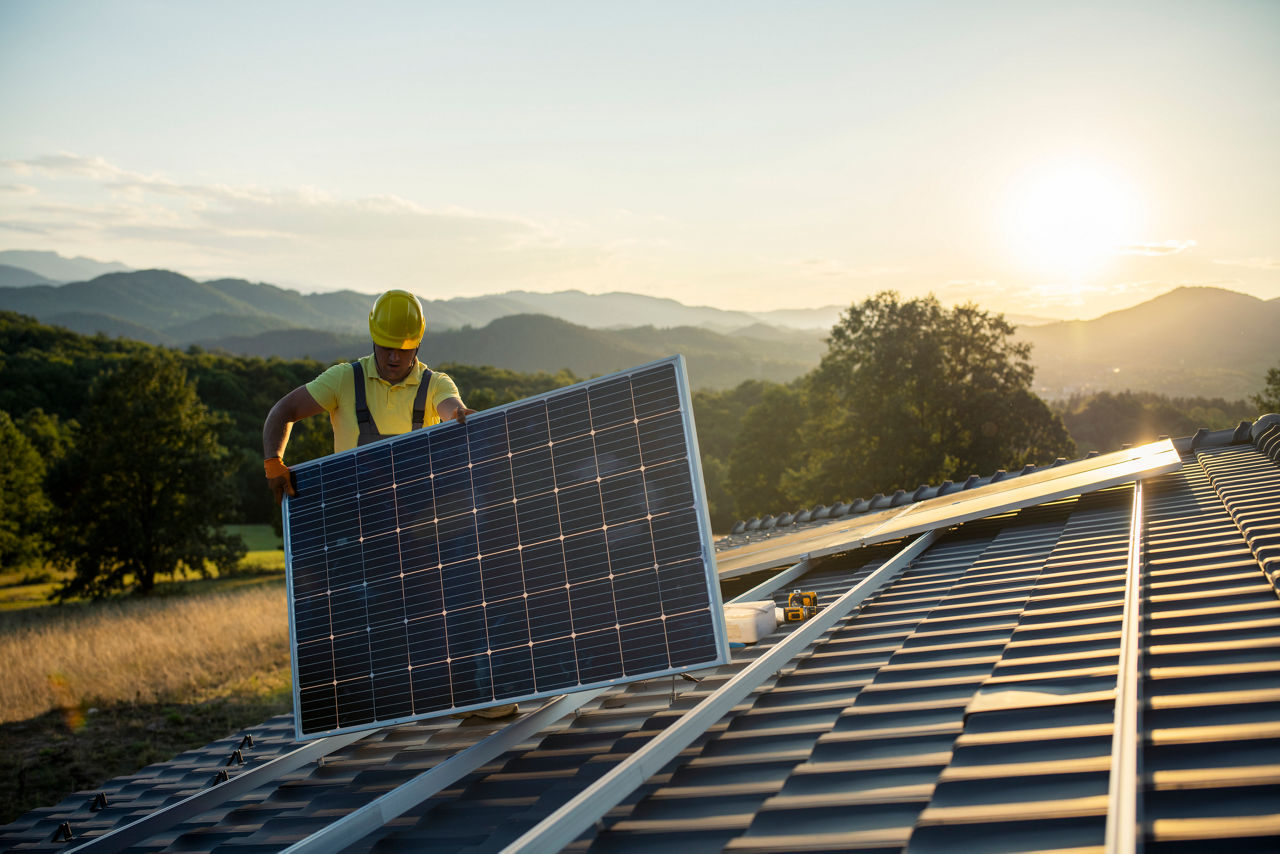 Arbeiter in Sicherheitskleidung installiert ein Solarpanel auf einem Hausdach bei Sonnenuntergang.