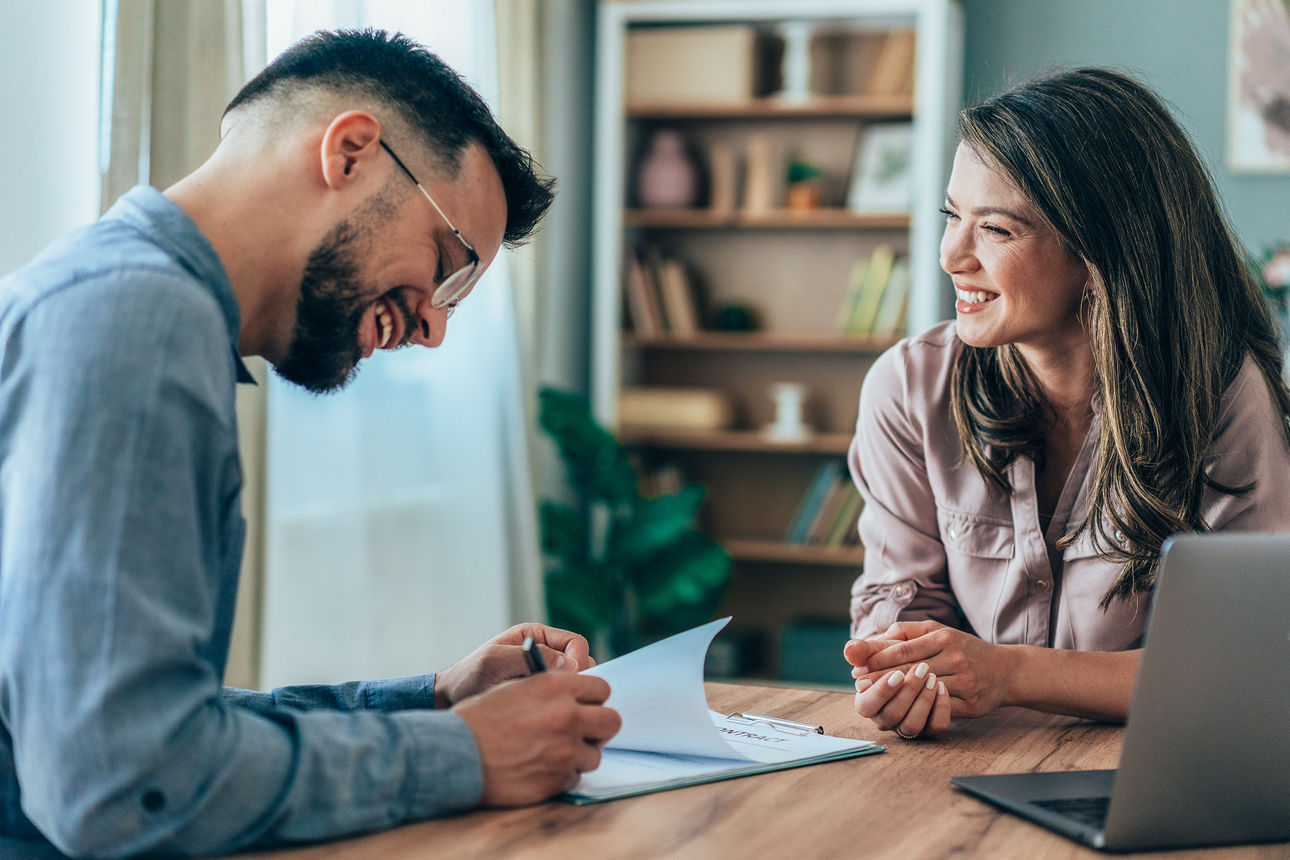 Eine Frau und ein Mann sitzen gegenüber am Tisch und der Mann unterschreibt gerade Unterlagen.