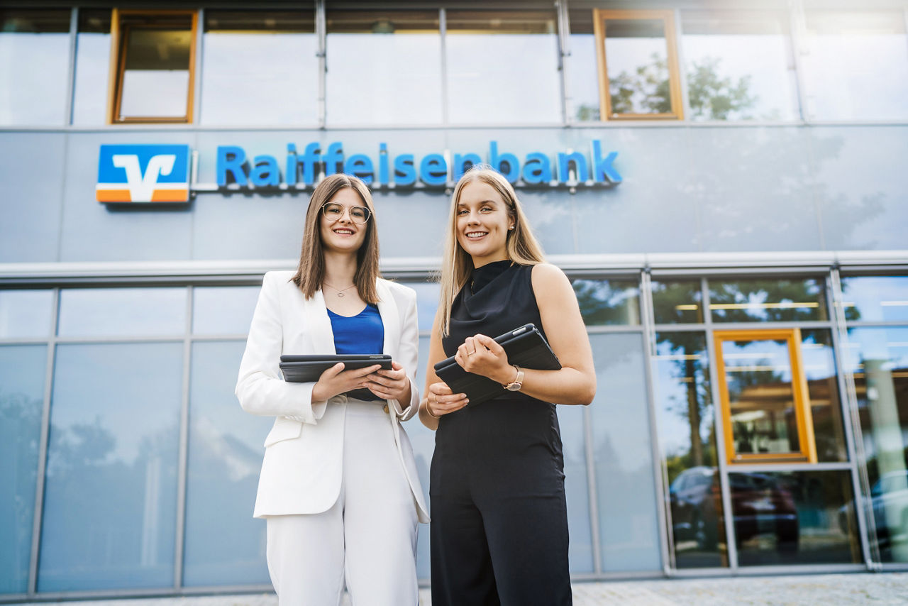 Zwei Frauen mit ihren Tablets vor einer Raiffeisenbank.