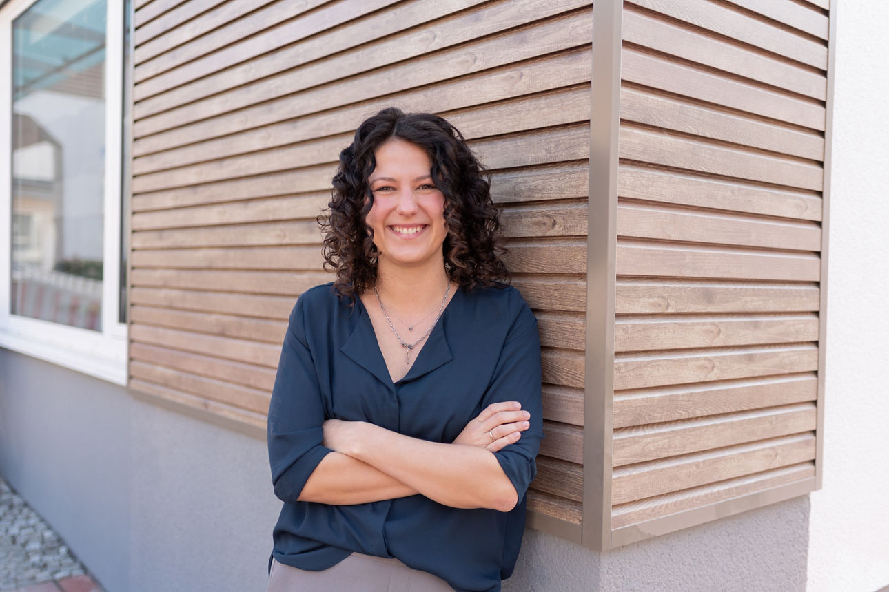 Junge Frau steht lächelnd mit verschränkten Armen an einer Hauswand