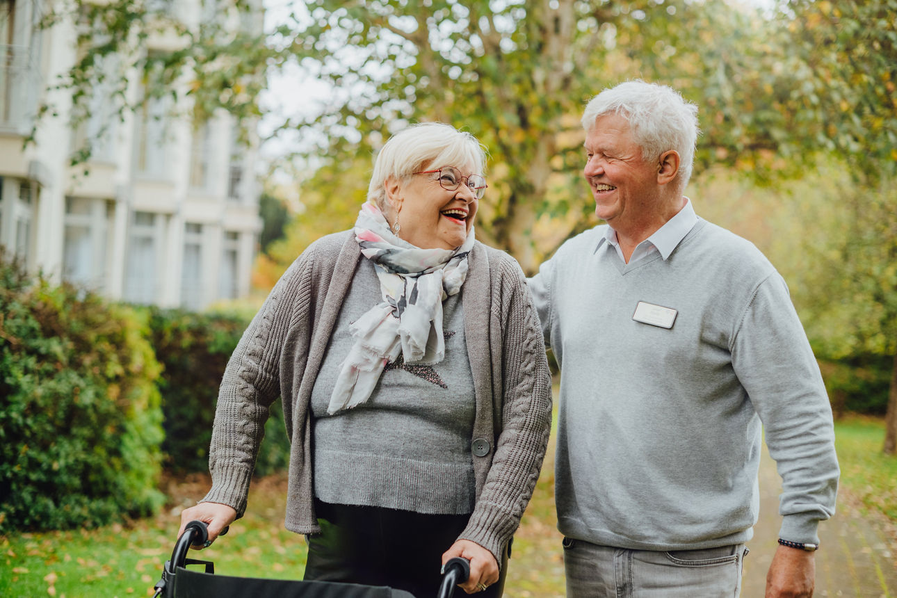 Ein Pfleger geht mit seiner Patienten, die am Rollator geht, im Park spazieren
