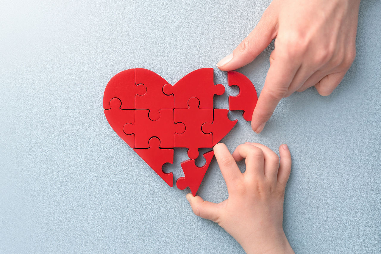 Eine Hand eines Erwachsenen und eine eines Kindes puzzeln ein rotes Herz