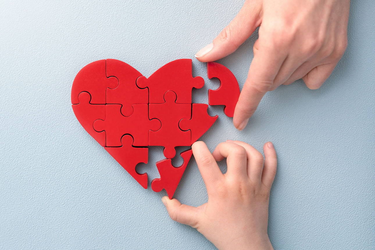 Eine Hand eines Erwachsenen und eine eines Kindes puzzeln ein rotes Herz