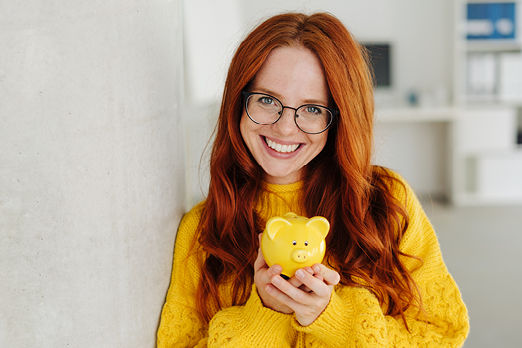 Lächelnde Frau mit Brille schaut in die Kamera und hat ein Sparschwein in der Hand.