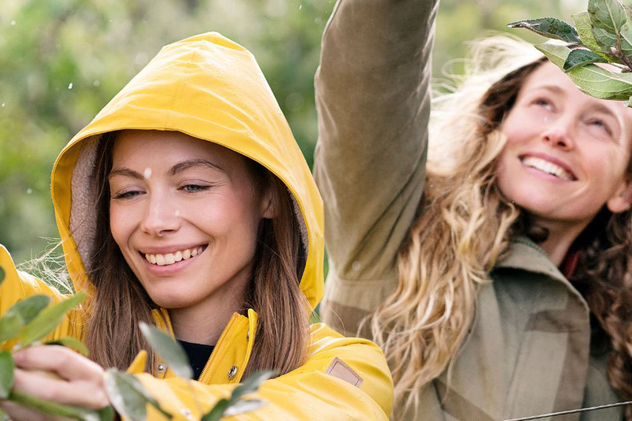 Eine Frau im gelben Regenmantel steht vor einem Obstbaum, während hinter ihr eine Frau Obst pflückt