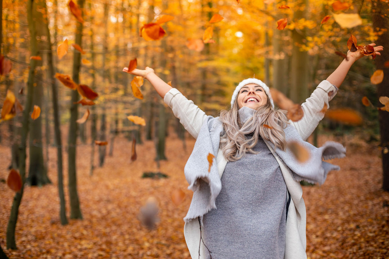 Eine Frau wirft in einem herbstlichen Wald Blätter in die Luft.