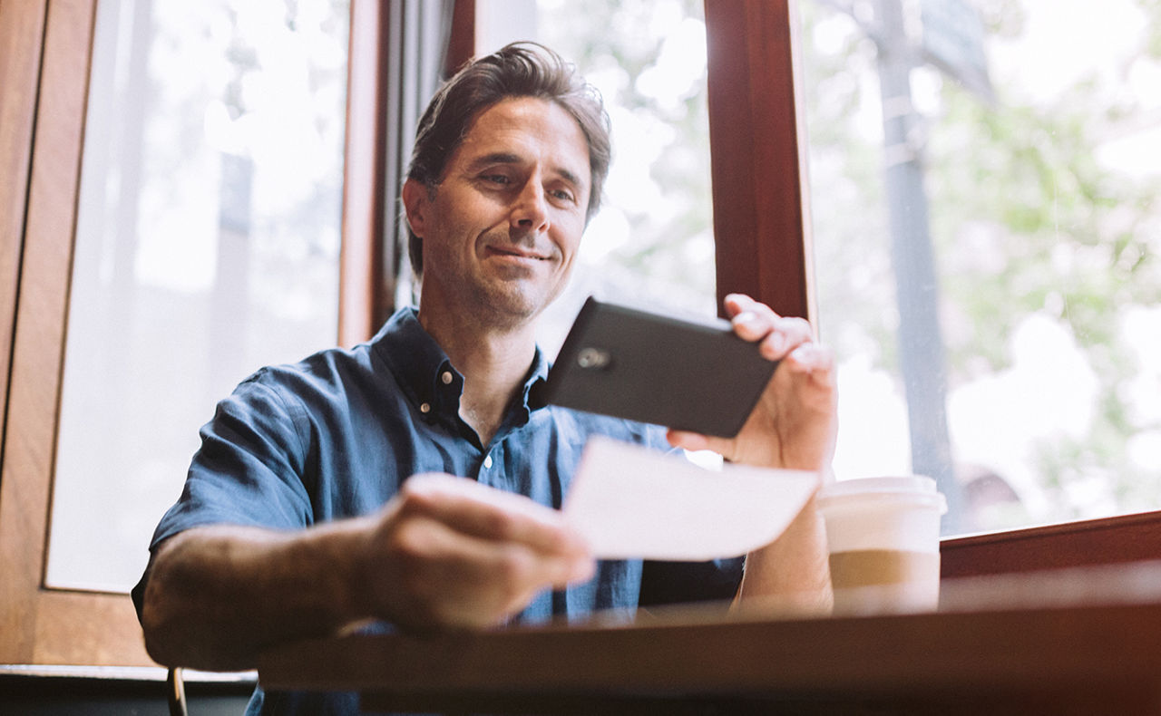 Ein Mann hält ein Smartphone in der einen Hand und ein Blatt Papier in der anderen Hand, das er fotografiert oder einscannt.