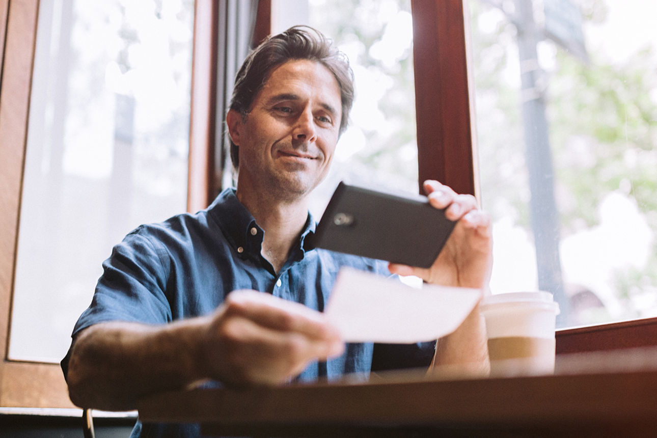 Ein Mann hält ein Smartphone in der einen Hand und ein Blatt Papier in der anderen Hand, das er fotografiert oder einscannt.