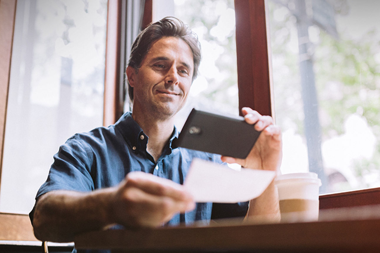 Ein Mann hält ein Smartphone in der einen Hand und ein Blatt Papier in der anderen Hand, das er fotografiert oder einscannt.