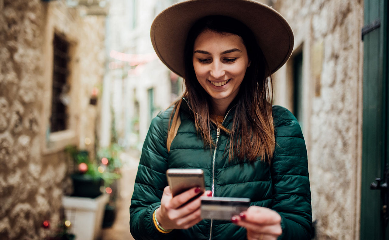 Frau steht in einer schmalen Gasse und hat ein Handy und eine girocard in der Hand