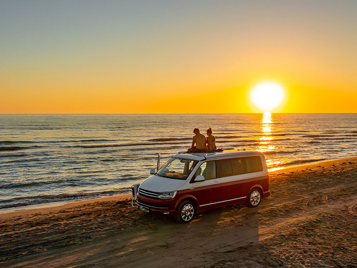 Zwei Personen stehen mit ihrem VW Bus an einem Strand bei Sonnenuntergang