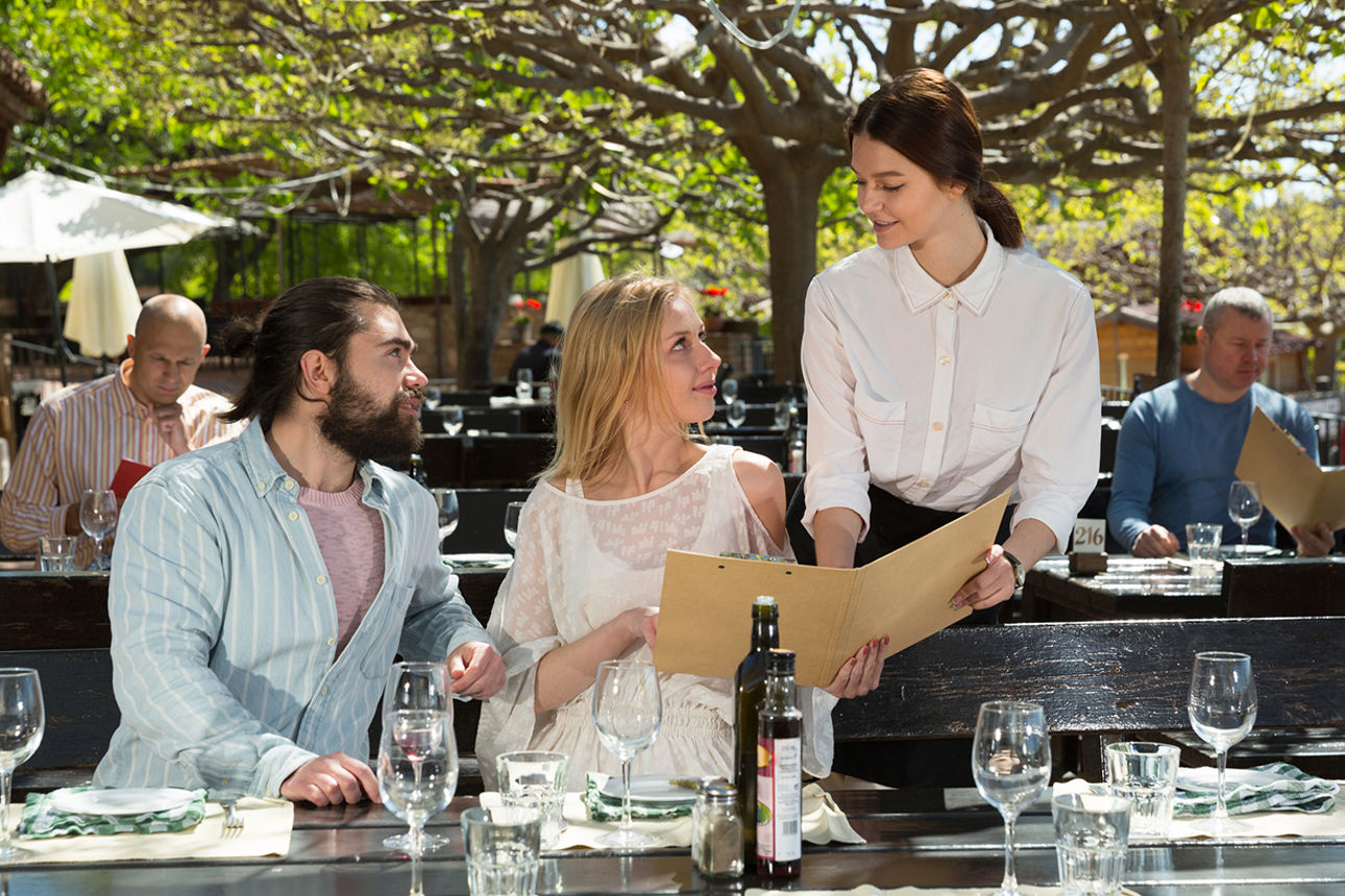 Ein Paar sitzt in einem Biergarten und lässt sich von einer Kellnerin die Karte zeigen