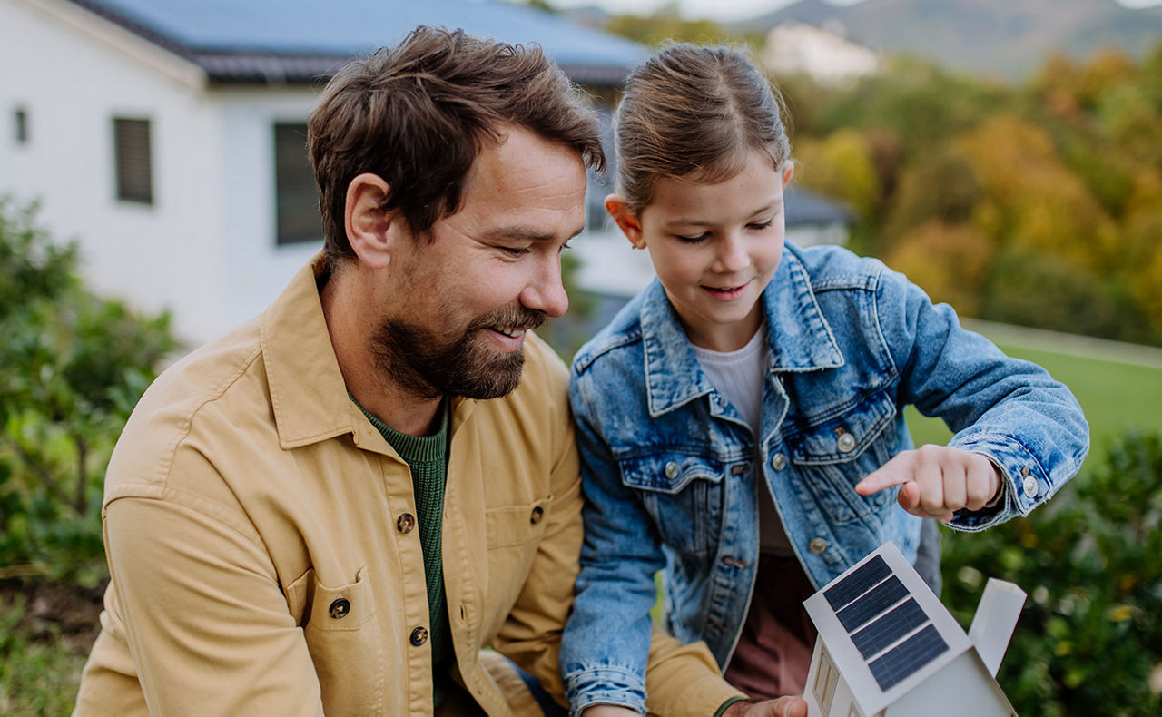 Mädchen mit ihrem Vater, das ein Papiermodell eines Hauses mit Sonnenkollektoren hält und Solarenergie erklärt