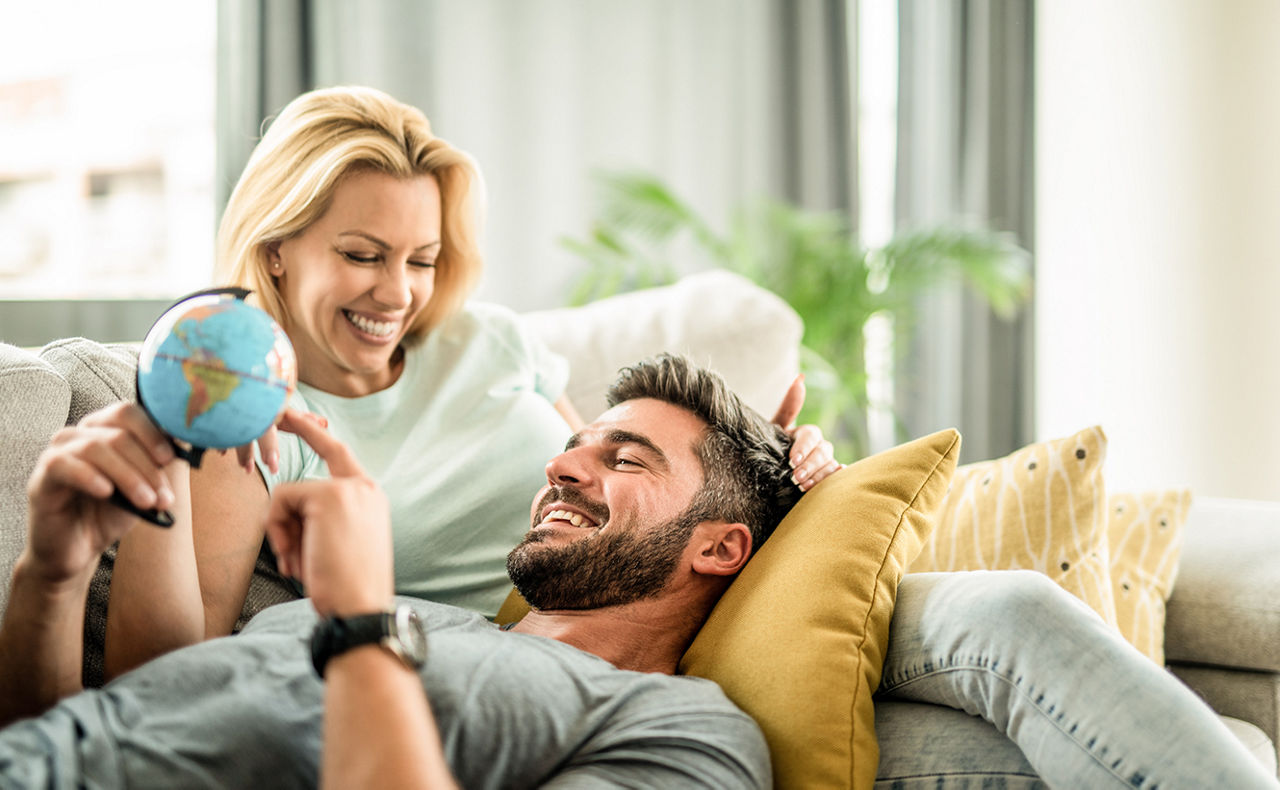 Mann und Frau liegen zusammen auf dem Sofa und schauen fröhlich nach einem Urlaubsziel auf einer Weltkugel
