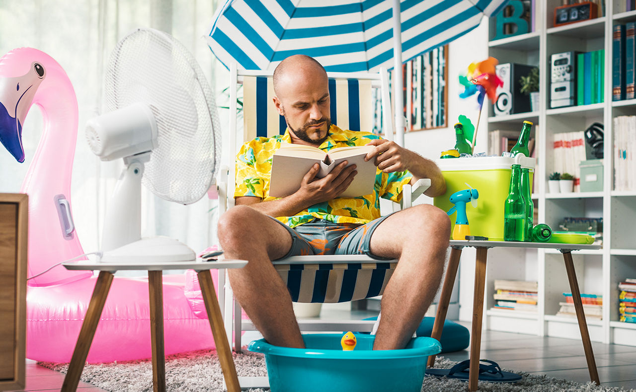 Kühle Wohnung an heißen Tagen – Mann sitzt mit Fußbad und Sonnenschirm im Wohnzimmer