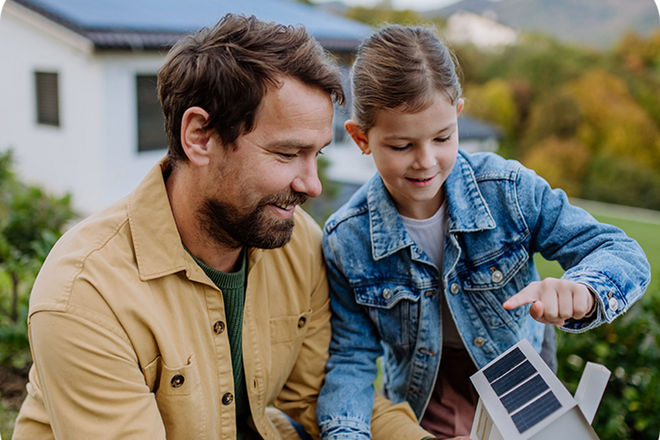 Mann hält mit seiner Tochter ein Miniatur-Haus mit Solarpanelen in der Hand