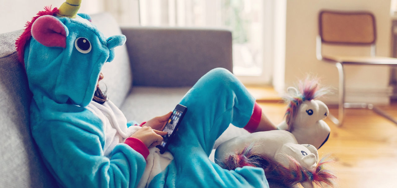 Kind im blauen Dino-Kostüm sitzt auf einem Sofa mit einem Handy in der Hand