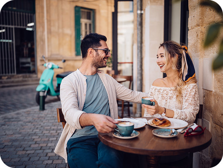 Ein Mann und eine Frau sitzen in einer Altstadt an einem Tisch und trinken Kaffee; im Hintergrund eine Vespa