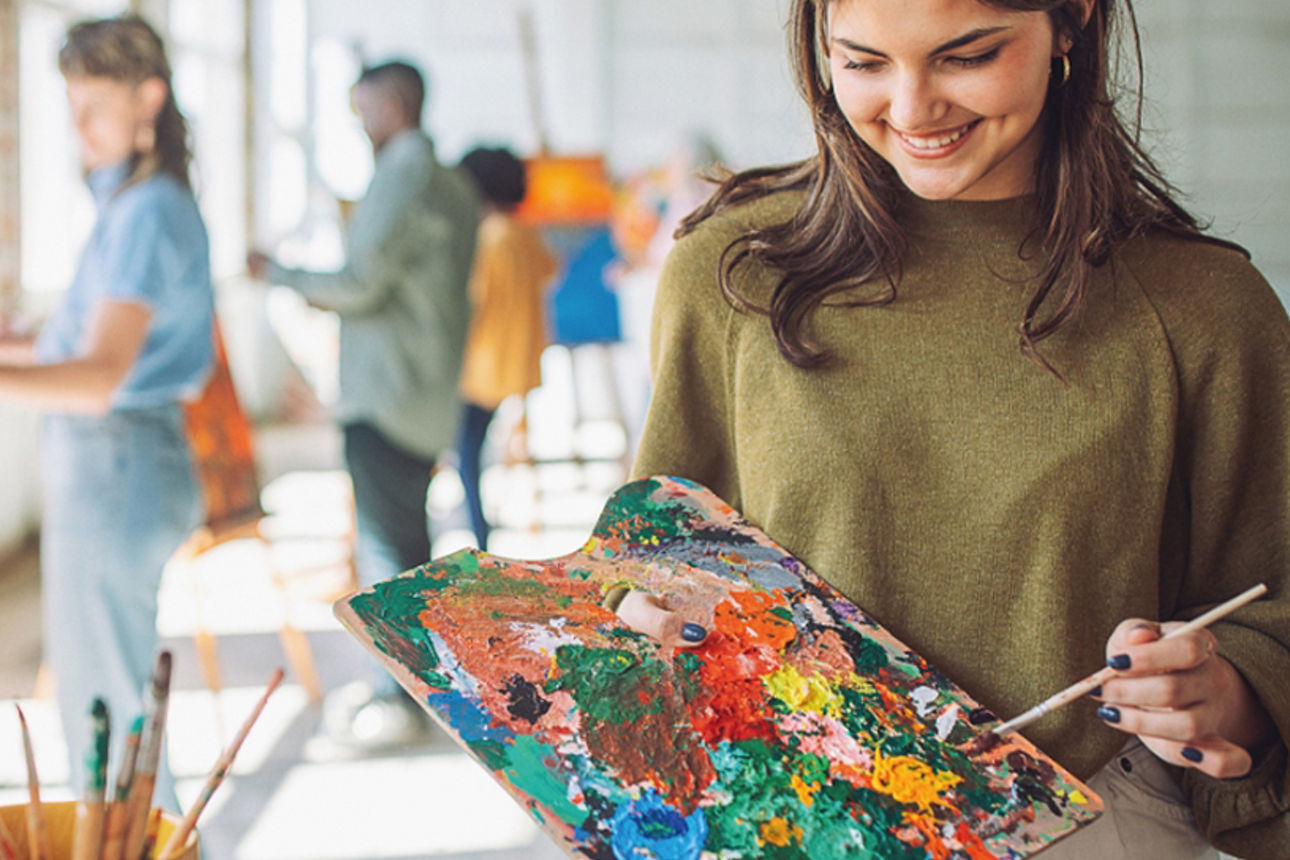 Eine junge Frau mit Farbpalette ist dabei, ein Bild zu malen. Im Hintergrund befinden sich weitere kreative, junge Menschen.