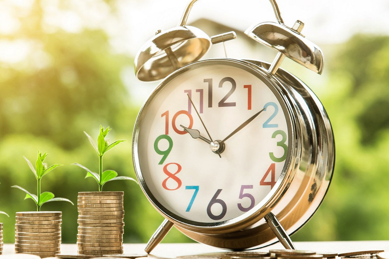 Drei kleine Stapel aus Münzen mit heraussprießenden Pflanzen und eine Uhr/Wecker