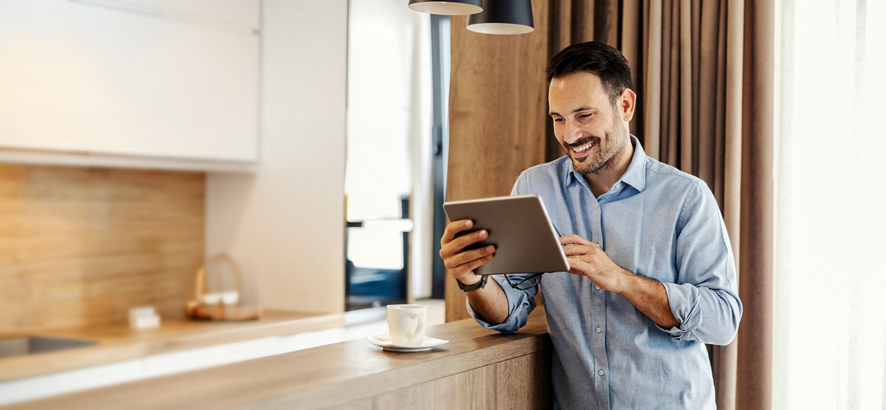 Junger Mann steht mit Tablet in der Hand in einer Küche und schaut mit einem Lachen im Gesicht auf ein Tablet, welches er in der Hand hält