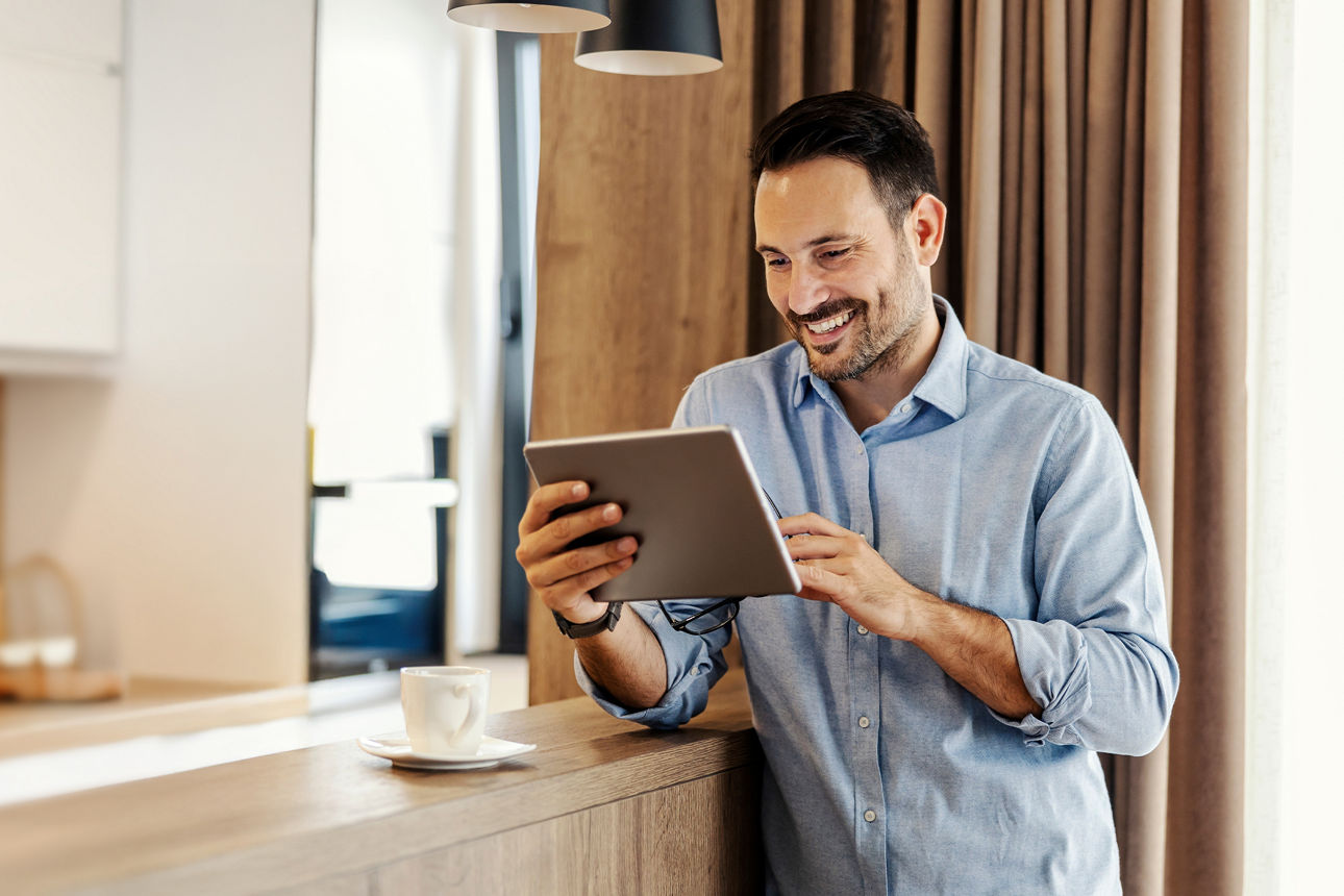 Junger Mann steht mit Tablet in der Hand in einer Küche und schaut mit einem Lachen im Gesicht auf ein Tablet, welches er in der Hand hält