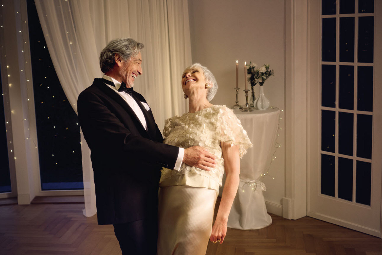 Eine elegant gekleidetes älteres Paar lacht und tanzt gemeinsam in einem festlich beleuchteten Raum.