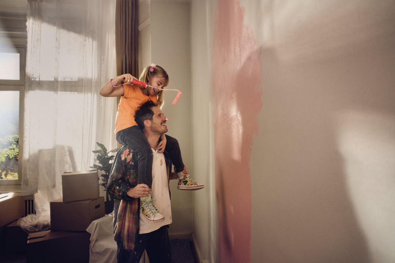 Ein Vater trägt seine Tochter auf den Schultern, während sie gemeinsam eine Zimmerwand mit Farbe streichen;