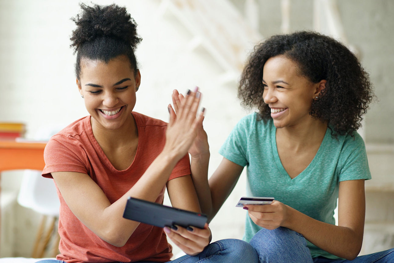 Zwei Frauen die lächelnd nebeneinander sitzen, ihre Smartphones in der Hand halten und sich abklatschen