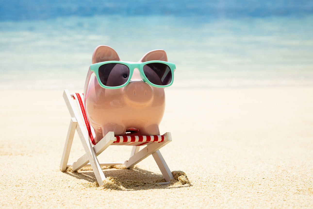 Ein Sparschwein mit Sonnebrille sitzt am Strand in einem Stuhl.