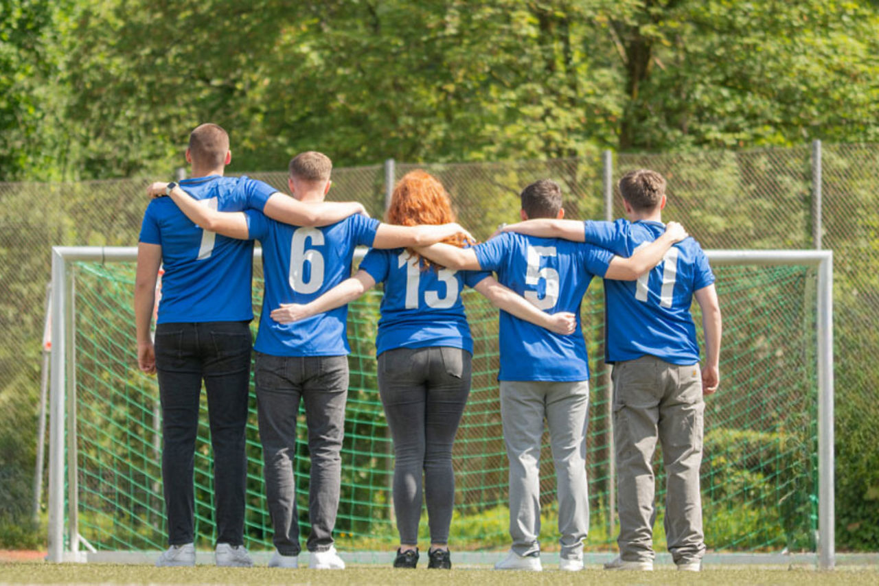 Fünf Personen stehen in blauen Trikots mit weißen Nummern auf dem Rücken vor einem Fußballtor. Sie haben die Arme umeinander 