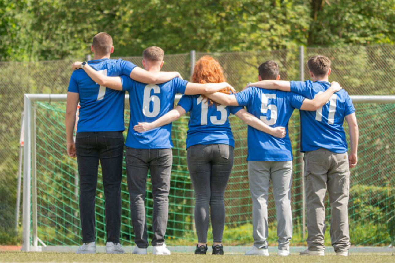 Fünf Personen stehen in blauen Trikots mit weißen Nummern auf dem Rücken vor einem Fußballtor. Sie haben die Arme umeinander 