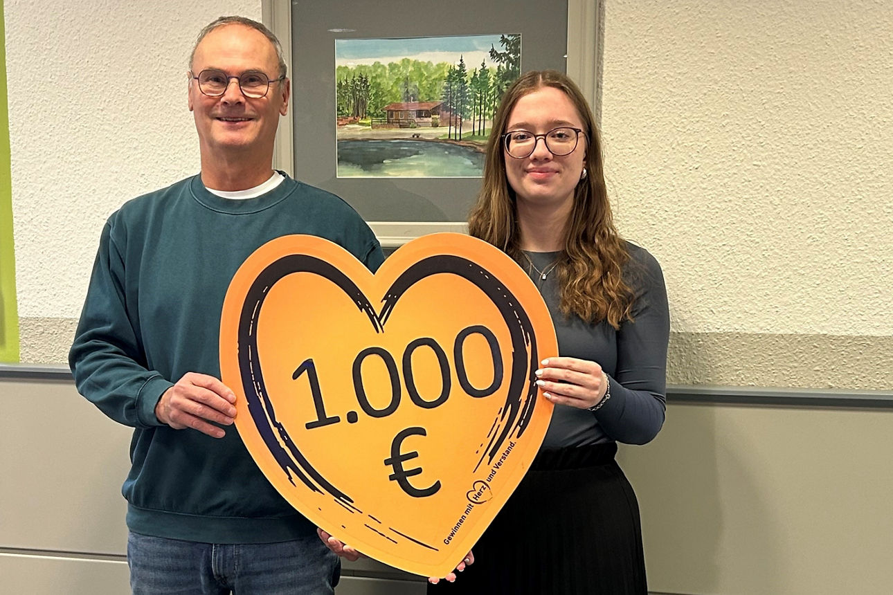 Der 2. Vorstand des Angelsportvereins und die Filialberaterin der VR-Bank stehen in einer Bankfiliale nebeneinander und halten ein Herz mit der Spendenaufschrift in den Händen. 