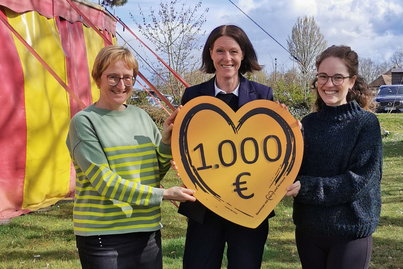 Es stehen drei Frauen bei Sonnenschein vor einem Zirkuszelt. Mittig steht die Regionalleiterin der VR-Bank. Sie hält ein Schild in Herzform mit der Spendenaufschrift in den Händen.