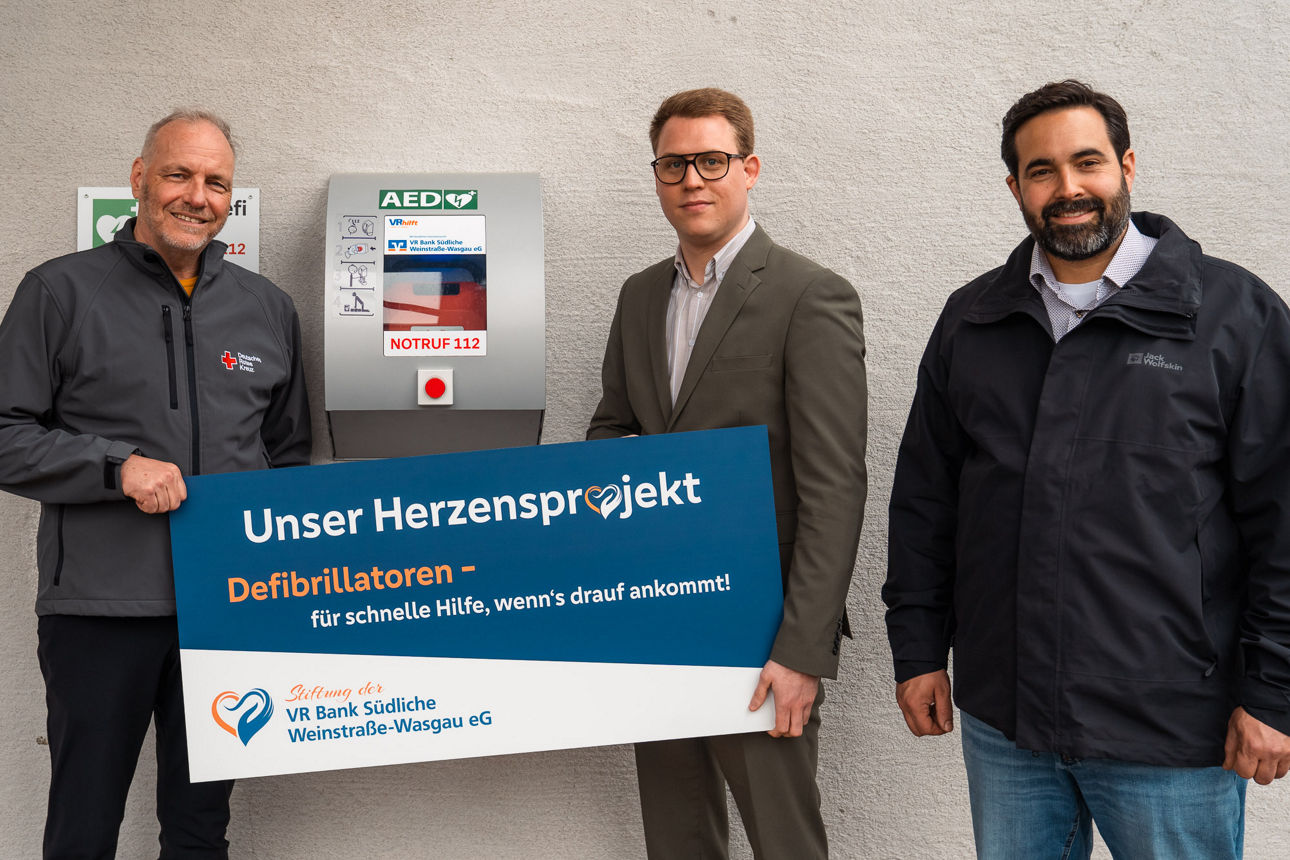 Drei Personen halten ein Banner zum Projekt ›Unser Herzensprojekt Defibrillatoren‹ vor einer Wand.