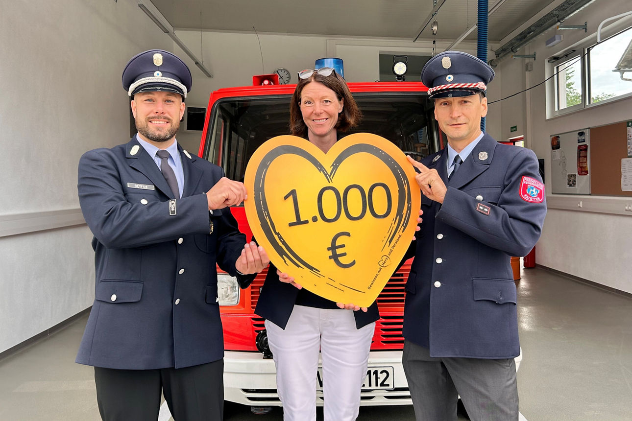 Vor dem Feuerwehrauto in einer Garage stehen die Regionalleiterin der VR-Bank, der Kommandant sowie der 1. Vorstand der Freiwilligen Feuerwehr. Sie halten ein Spendenherz mit Aufschrift der Spendensumme in den Händen.