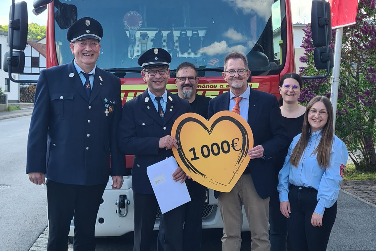Es stehen fünf Mitglieder der Freiwilligen Feuerwehr Schönau, zwei davon in Uniform, mit dem Spendenbotschafter der Bank, vor einem Feuerwehrfahrzeug. Sie halten ein Schild in Herzform mit Aufschrift der Spendensumme in den Händen. 