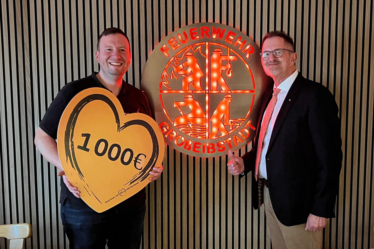 Auf dem Bild stehen der 1. Vorsitzende des Feuerwehrvereins Großeibstadt und der Spendenbotschafter der VR-Bank. Sie stehen vor einem beleuchteten Wappen der Feuerwehr und halten ein Schild in Herzform mit der Spendenaufschrift in den Händen. 