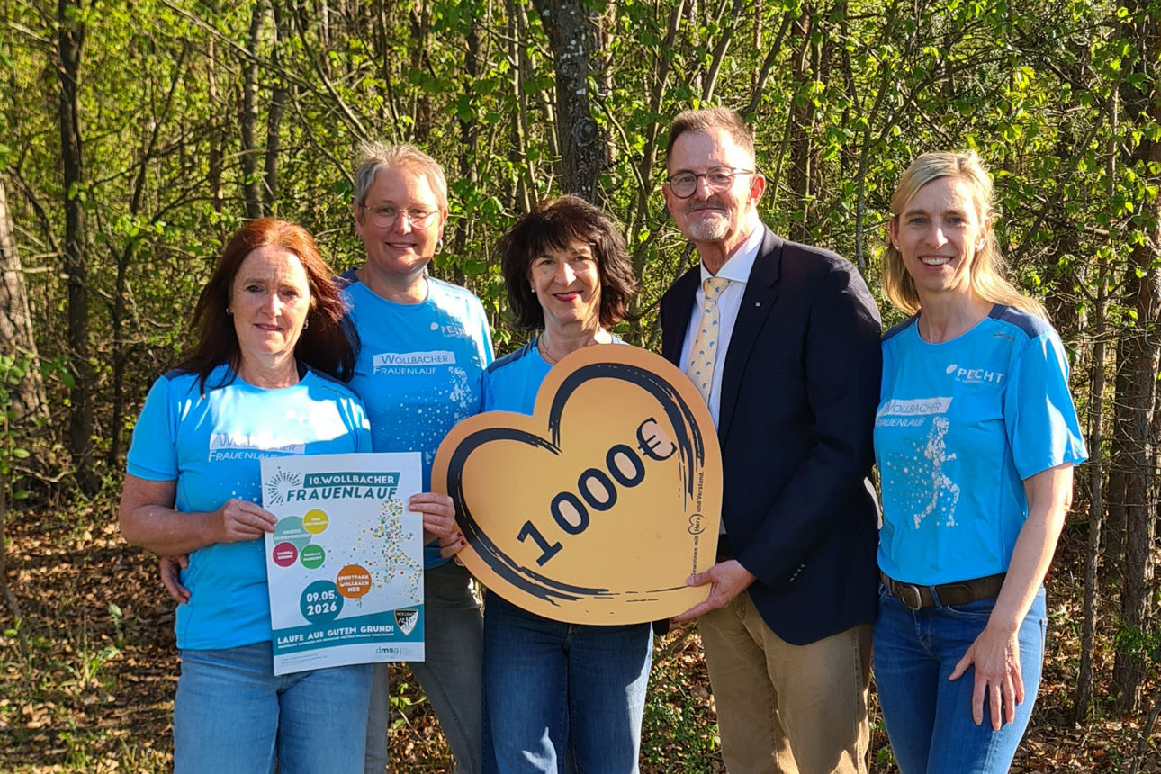 Das Foto wurde im Freien bei Sonnenschein aufgenommen. Es zeigt die Verantwortlichen des Wollbacher Frauenlaufes und den Spendenbotschafter der Bank. Sie halten ein Spendenherz mit Aufschrift der Spendensumme in den Händen. 