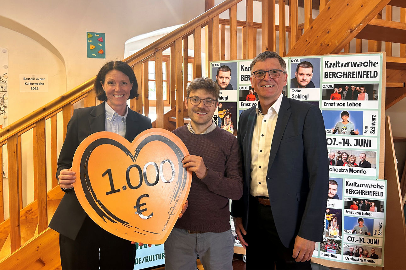 Auf dem Bild stehen in einem Raum vor einer Treppe, die Regionalleiterin der VR-Bank, der Bürgermeister Bergrheinfeld und ein Mitglied der Gemeindeverwaltung. Sie halten ein Schild in Herzform mit Aufschrift der Spendensumme in den Händen.
