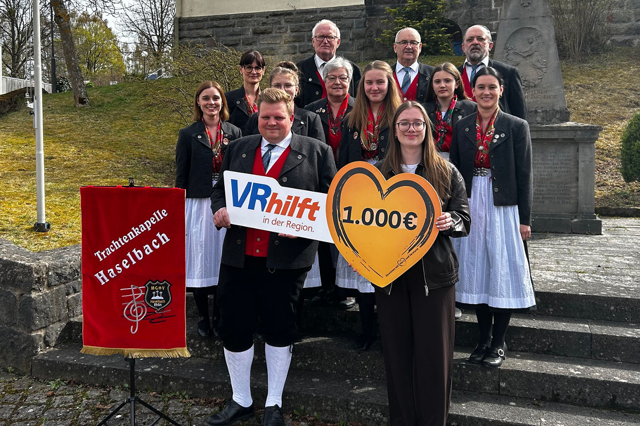 Auf dem Bild stehen außen auf einer Treppe die Mitglieder des Musikvereins Haselbach in ihrer Tracht, sowie die Filialberaterin der VR-Bank.  Sie hält ein Schild in Herzform in den Händen, mit der Aufschrift der Spendensumme. 
