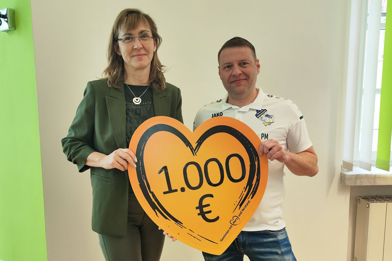 In der Bankfiliale stehen die Filialberaterin der Bank und der 1. Vorsitzende des Vereins. Sie lächeln in die Kamera und halten das Spendenherz mit Aufschrift der Spendensumme in den Händen. 