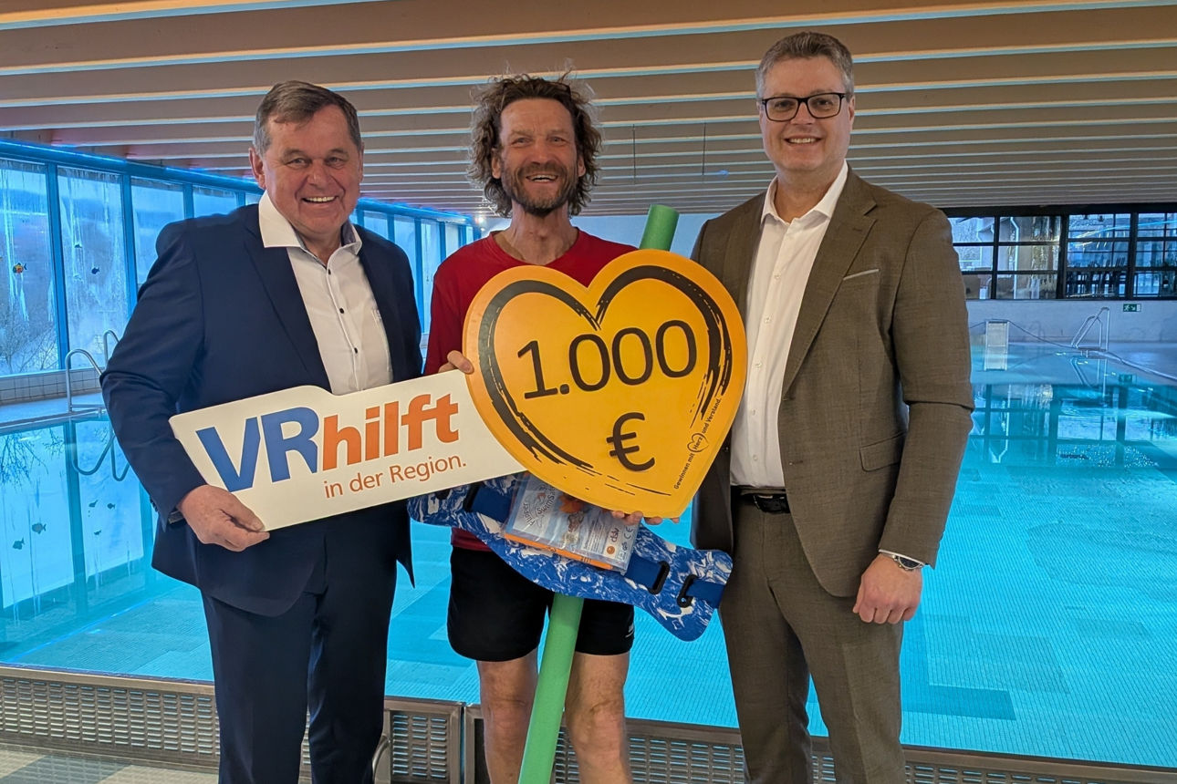 Das Bild wurde im Hallenbad aufgenommen, es stehen der Bürgermeister der Stadt Arnstein, der Schwimmmeister des Hallenbades und der Vorstand der VR-Bank vor einem Schwimmbecken.  Sie halten ein Herz in den Händen mit Aufschrift der Spendensumme. 