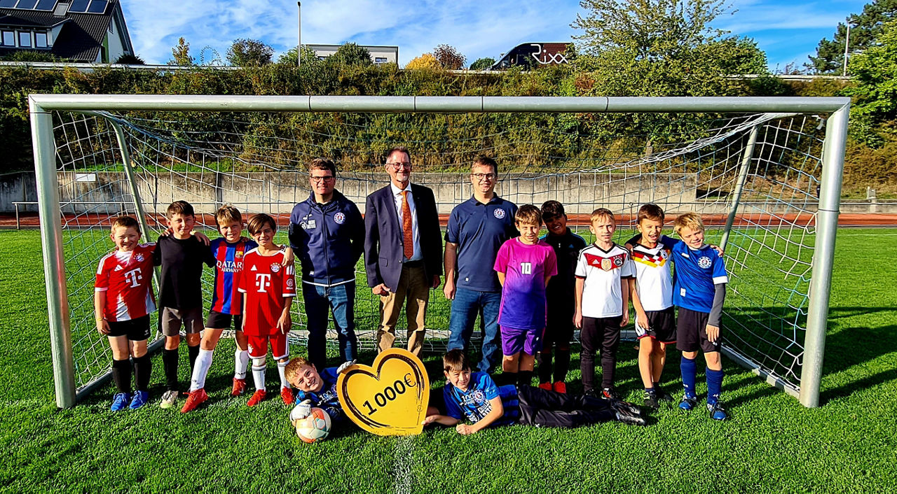 Auf dem Bild sind die Nachwuchskicker des TSV Wülferhausen mit dem Spendenbotschafter der VR-Bank und dem Vorstand des Vereins zu sehen. Sie stehen in einem Fußballtor, die Sonne scheint.