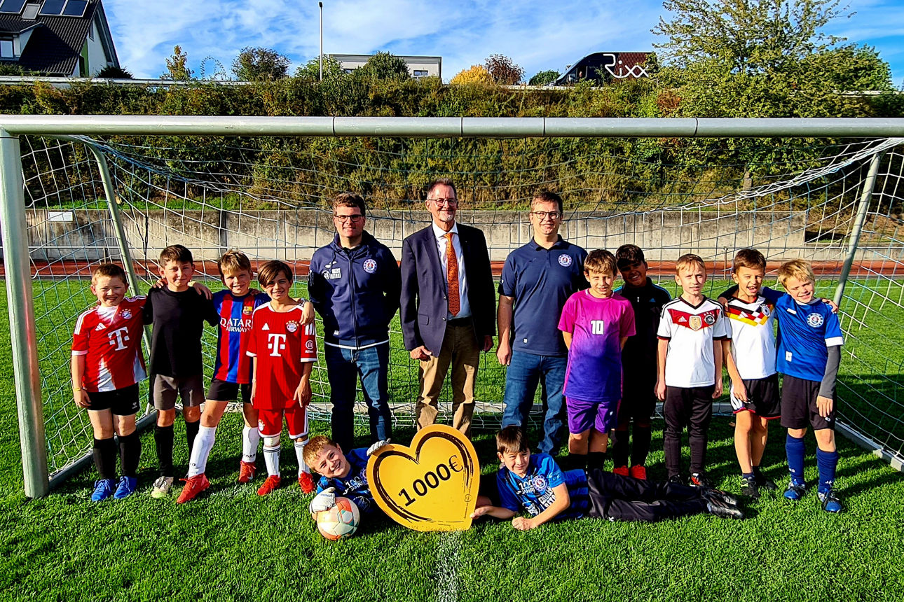 Auf dem Bild sind die Nachwuchskicker des TSV Wülferhausen mit dem Spendenbotschafter der VR-Bank und dem Vorstand des Vereins zu sehen. Sie stehen in einem Fußballtor, die Sonne scheint.