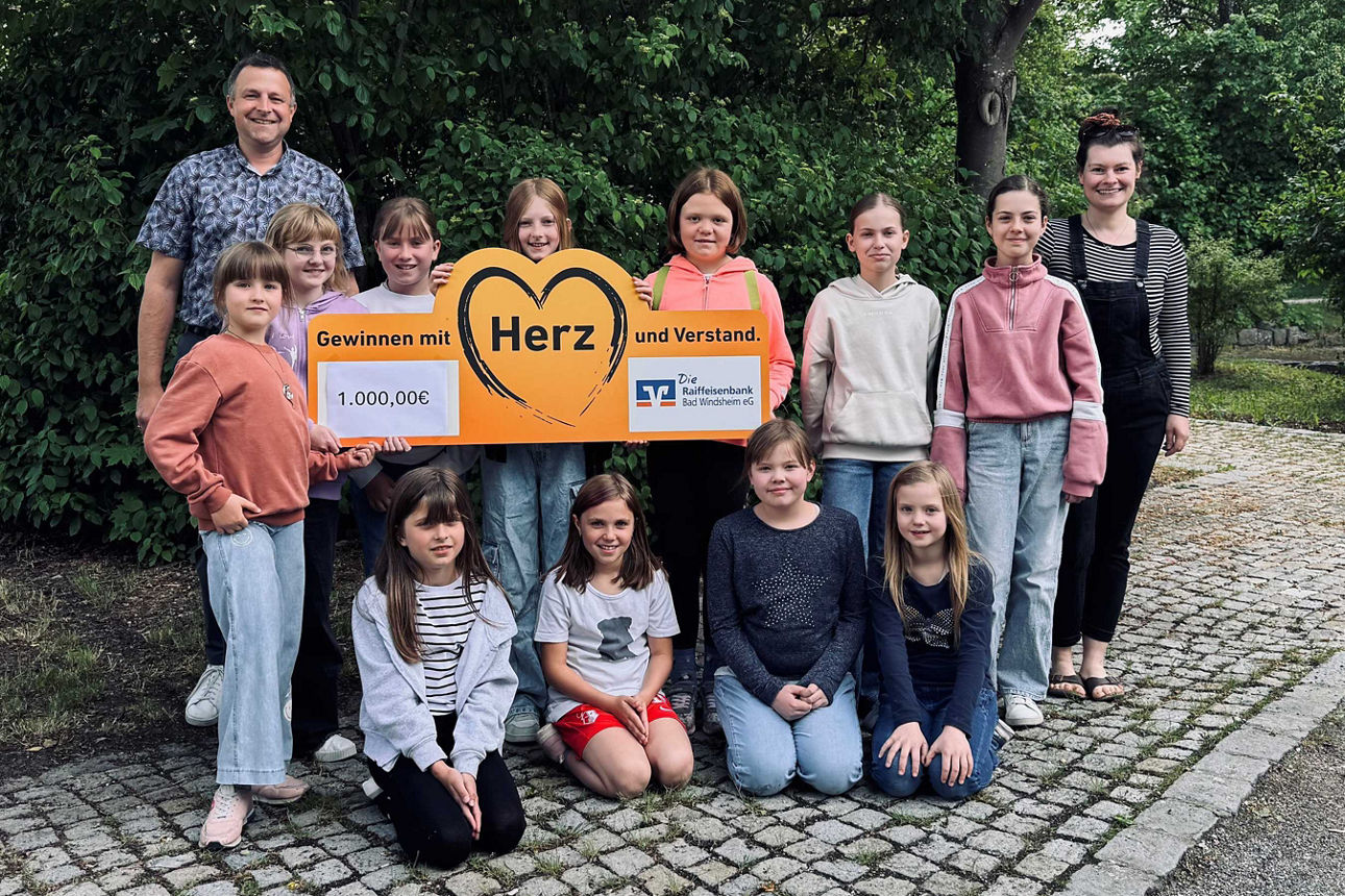 Gruppenbild mit elf Mädchen halten ein Schild mit Herz und Aufschrift von 1.000 Euro Spende hinten links steht ein Mann und rechts eine Frau. Alle lächeln in die Kamera.