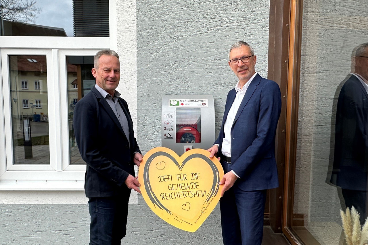 Zwei Männer halten ein großes gelbes Herz mit der Aufschrift „DEFI FÜR DIE GEMEINDE REICHERTSHEIM“ vor einem an der Wand mont