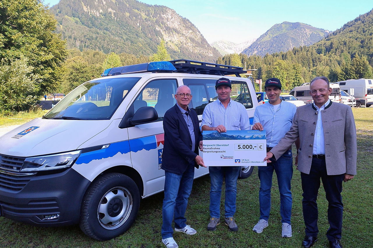 Spende an Bergwacht Oberstdorf - 4 Personen mit Scheck, im Hintergrund die Oberstdorfer Berge und das Einsatzfahrzeug der Bergwacht