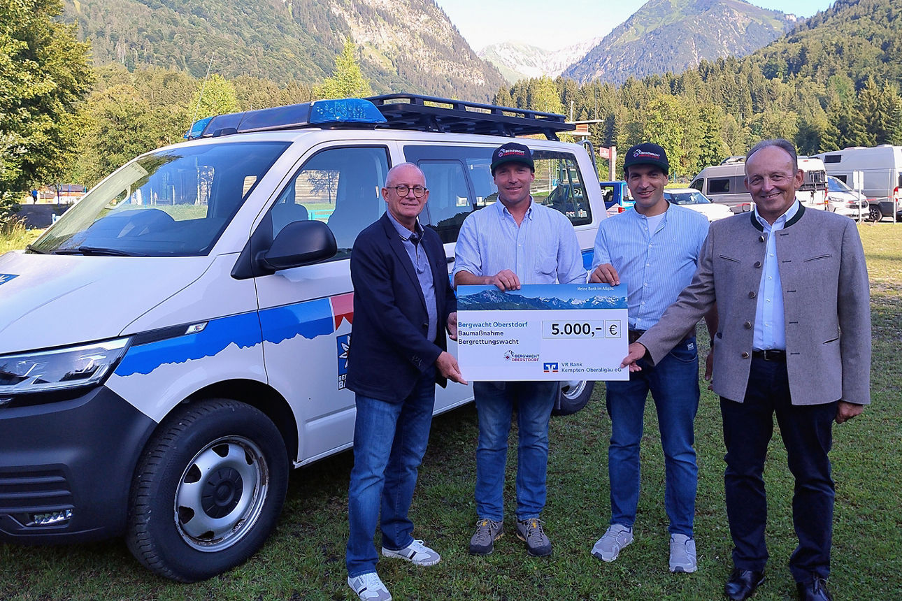 Spende an Bergwacht Oberstdorf - 4 Personen mit Scheck, im Hintergrund die Oberstdorfer Berge und das Einsatzfahrzeug der Bergwacht