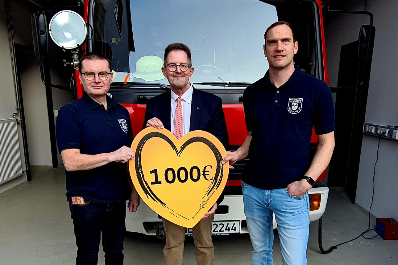 Es stehen der Spendenbotschafter der VR-Bank und der ehemalige und aktuelle Kommandant der Feuerwehr Hausen vor dem Feuerwehrfahrzeug und halten ein Herz mit Aufschrift der Spendensumme in den Händen. 
